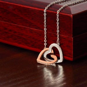 May include: A silver necklace with two interlocking heart pendants. One heart is rose gold with clear crystals, the other is silver with clear crystals. The necklace is displayed on a dark red wooden surface.