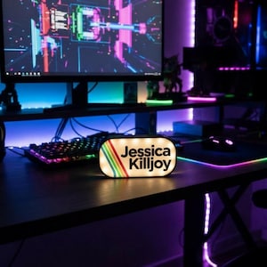 May include: A lighted sign with the name "Jessica Killjoy" in a retro font, featuring a rainbow stripe design. The sign is on a desk with a computer, keyboard, and mouse, illuminated by colorful LED lights.