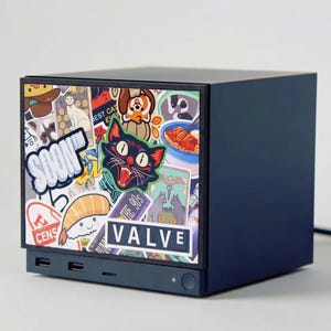 May include: A dark blue rectangular computer case featuring a front panel covered in various colorful stickers, including a black cat, a sushi roll, and a prominent white rectangular sticker with the black text VALVE and the word SOON.