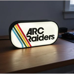 May include: A rectangular, illuminated sign with the text "ARC Raiders" in bold, black letters. The sign has a white background with a colorful stripe design of red, yellow, green, and blue. The sign is on a wooden desk.