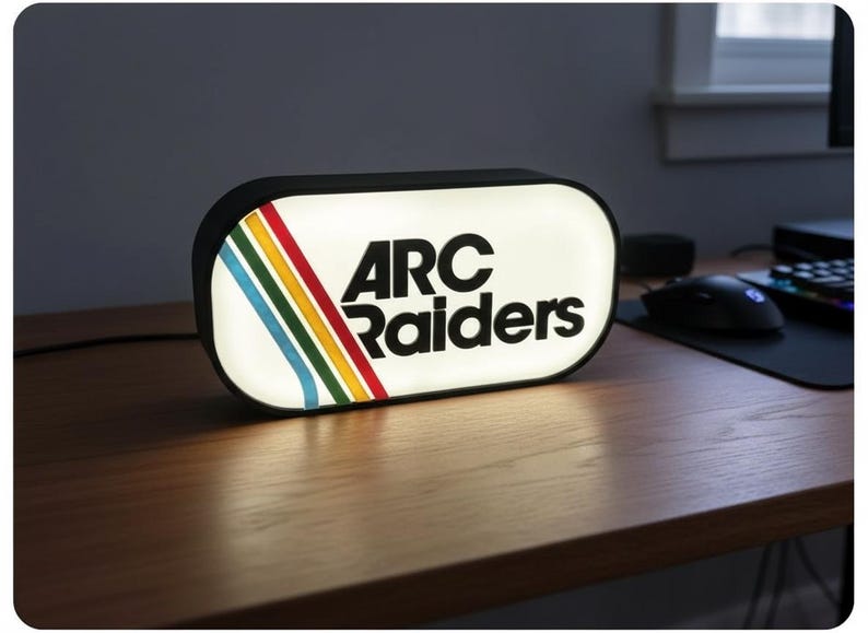May include: A rectangular, illuminated sign with the text "ARC Raiders" in bold, black letters. The sign has a white background with a colorful stripe design of red, yellow, green, and blue. The sign is on a wooden desk.