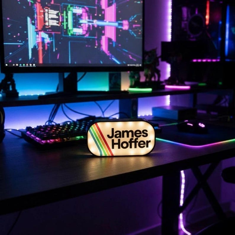 May include: A lighted, rectangular sign with the name "James Hoffer" in white text. The sign has a retro design with rainbow stripes and is on a desk with a keyboard and monitor in the background. The room is lit with colorful LED lights.