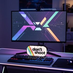 May include: A gaming setup featuring a curved monitor displaying a colorful geometric design. A keyboard, mouse, and a sign that reads "don't shoot" are on the desk. The setup is illuminated with neon lights.