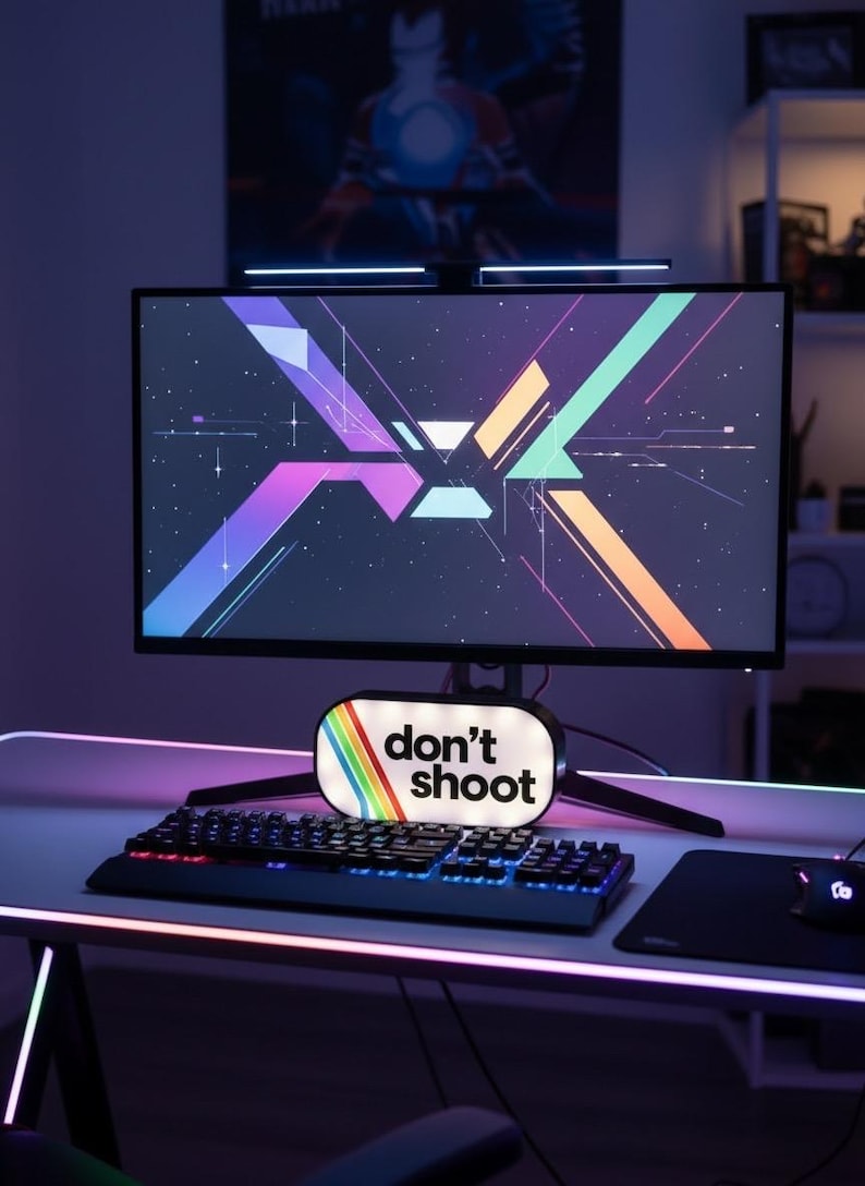 May include: A gaming setup featuring a curved monitor displaying a colorful geometric design. A keyboard, mouse, and a sign that reads "don't shoot" are on the desk. The setup is illuminated with neon lights.