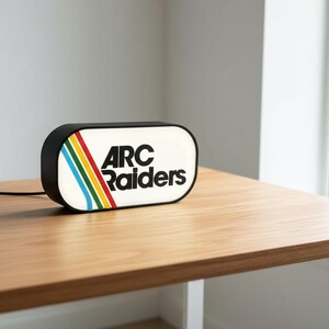 May include: A rectangular black and white light-up sign with the text "ARC Raiders" in black. The sign has a retro design with colorful stripes. It sits on a wooden table.