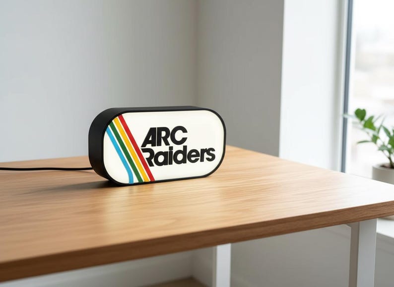 May include: A rectangular black and white light-up sign with the text "ARC Raiders" in black. The sign has a retro design with colorful stripes. It sits on a wooden table.