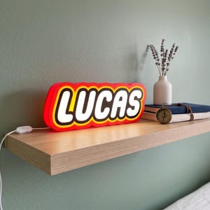 May include: A custom illuminated sign featuring the name LUCAS in bold white capital letters, surrounded by a thick red and yellow border. The sign is rectangular with rounded edges and includes a white power cord with an inline switch.