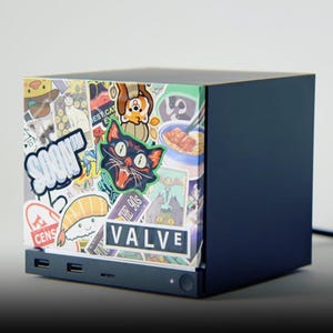 May include: A dark blue cube-shaped computer console featuring a front panel covered in various colorful stickers, including a cat, sushi, and the text "SOON" and "VALVE". The device has visible USB ports and a power button on the lower front edge.