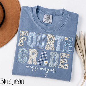 Peut inclure: Un t-shirt bleu jean avec un col rond et une étiquette de marque Comfort Colors. Le devant présente le texte "FOURTH GRADE" en grandes lettres de style bloc avec divers motifs floraux et vichy bleus et blancs, ainsi que "miss mayor" en écriture blanche.