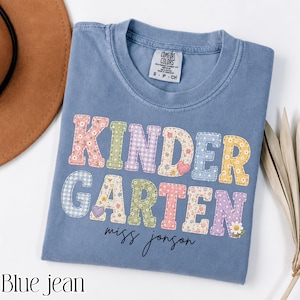 Può includere: Una t-shirt color blu jeans con la parola KINDERGARTEN in grandi lettere maiuscole dai motivi vari e colori pastello, inclusi fiori, quadretti vichy e pois. Sotto la parola, il nome Miss Jonson è stampato in un carattere corsivo nero.