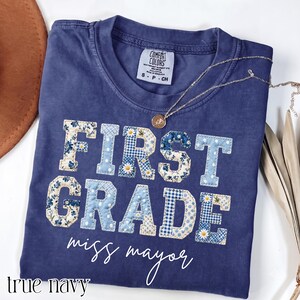 Puede incluir: Una camiseta azul marino con el texto FIRST GRADE en letras grandes de estilo bloque con un patrón de patchwork de telas con flores azules, cuadros vichy y lunares. Debajo del texto, las palabras miss mayor están en cursiva blanca.
