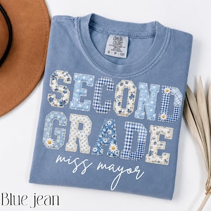 Puede incluir: Una camiseta azul de manga corta con el texto SECOND GRADE en grandes letras de tela aplicada con varios patrones florales y de cuadros vichy en azul y blanco. El texto miss mayor está impreso en cursiva blanca bajo las letras.