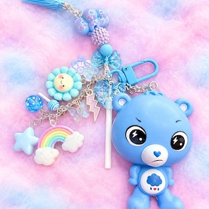 May include: A blue bear keychain featuring a grumpy expression and a rain cloud belly badge. The keychain includes a silver chain with charms including a smiling flower, a rainbow, a star, a lightning bolt, a butterfly, and a blue tassel.