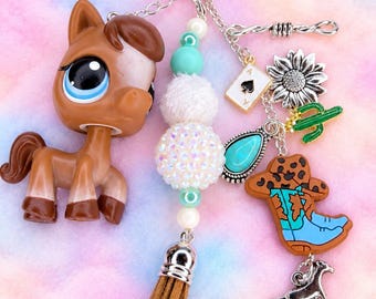 May include: A keychain featuring a brown horse figurine with large eyes, a tan suede tassel, and a strand of white and turquoise beads. Attached charms include a silver star, an ace of spades card, a sunflower, a green cactus, a cowboy boot, and a horse.