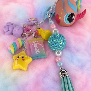 May include: A keychain featuring a peach-colored fish figurine, a turquoise suede tassel, and various charms including a yellow star, a yellow duck, a purple bear, a multi-colored candy, and a rectangular charm with the word Sweet printed on it.