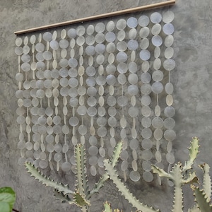 Puede incluir: Un adorno de pared decorativo hecho de numerosas piezas pequeñas, iridiscentes, de forma circular y ovalada. Las piezas están ensartadas y suspendidas de una barra de madera. El fondo es una pared de hormigón gris, y un cactus es visible.