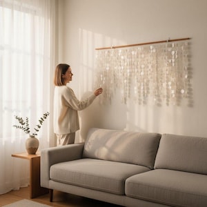 Puede incluir: Una decoración de pared con numerosos elementos pequeños, redondos, blanquecinos, similares a conchas, suspendidos de una barra de madera. La decoración está montada en una pared blanca sobre un sofá gris.