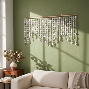 May include: A wall hanging composed of numerous circular, translucent white shell discs suspended from a horizontal wooden rod. The discs are arranged in multiple vertical strands of varying lengths, creating a layered, cascading effect.