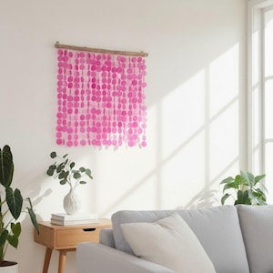 Könnte beinhalten: Ein Wandbehang bestehend aus mehreren vertikalen Strängen aus kreisförmigen, durchscheinenden, leuchtend pinkfarbenen Scheiben, die an einer horizontalen Holzstange hängen. Die Scheiben sind gitterartig angeordnet und erzeugen einen lebendigen Effekt.