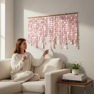 May include: A wall hanging composed of numerous light pink circular discs suspended from a horizontal wooden rod. The discs are arranged in vertical strands of varying lengths, creating a layered, cascading effect across the wall surface.