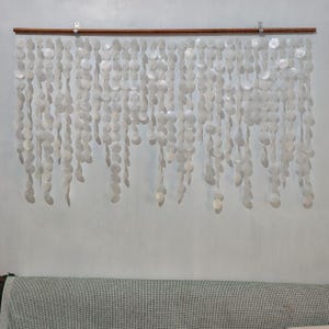May include: A wall hanging composed of numerous white, circular, shell-like discs suspended from a wooden rod. The discs are arranged in vertical strands, creating a cascading effect. The wall hanging is mounted on a light-coloured wall.