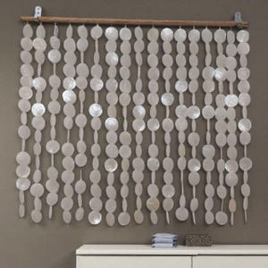 May include: A wall hanging consisting of multiple vertical strands of circular, iridescent white shell discs suspended from a horizontal wooden rod. The strands feature alternating sizes of round, flat shell pieces that create a textured, layered appearance across the wall surface.