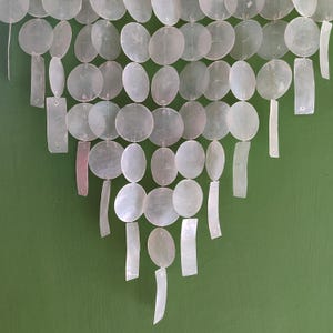 May include: A decorative wall hanging composed of iridescent, oval and rectangular shell pieces. The shell pieces are strung together, creating a cascading effect. The background is a solid, muted green.