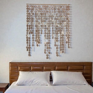 Könnte beinhalten: Ein dekorativer Wandschmuck aus zahlreichen kleinen, runden, metallisch getönten Scheiben, die an einer Holzstange aufgehängt sind. Die Scheiben hängen in vertikalen Strängen und erzeugen einen Kaskadeneffekt. Über einem Bett mit braunem Lederkopfteil und weißen Kissen.