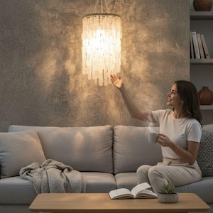 May include: A modern, cylindrical chandelier with a white, layered design hangs above a living room. The light fixture is suspended from the ceiling. A woman sits on a gray sofa, holding a mug. A wooden coffee table and a blanket are in the foreground.