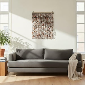 Könnte beinhalten: Ein anthrazitfarbenes Sofa mit einer hellbeigen Decke, die über eine Armlehne drapiert ist. Über dem Sofa hängt ein Wandbehang aus zahlreichen kleinen, runden, metallisch glänzenden Scheiben, die an einer horizontalen Holzstange hängen.