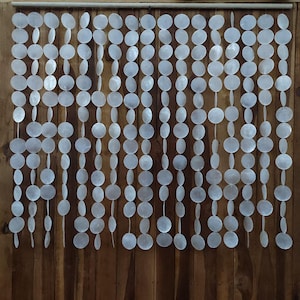 Puede incluir: Una cortina decorativa hecha de piezas de concha blancas, iridiscentes, circulares y ovaladas ensartadas. La cortina cuelga de una barra de madera sobre un fondo de madera marrón. Las piezas de concha son de varios tamaños y formas.