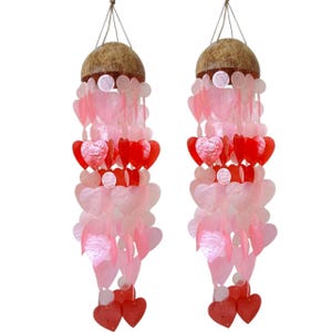 May include: Two heart-shaped wind chimes. Each chime features a coconut top, strings of pink and red heart-shaped shells, and a few white circular accents. The chimes are suspended by a thin, natural-colored string.