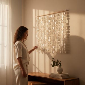 May include: A wall hanging composed of multiple vertical strands of circular, translucent white shell discs suspended from a horizontal wooden rod. The discs are arranged in a grid pattern, creating a light, layered effect against the wall.