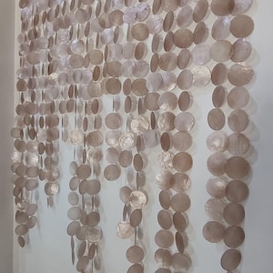 Op de afbeelding: Een decoratieve wanddecoratie gemaakt van talrijke, ronde, iriserende, schelpachtige schijven. De schijven zijn in verticale strengen geregen, waardoor een gordijnachtig effect ontstaat. Het kleurenpalet is voornamelijk neutraal, met tinten beige en lichtbruin, wat een subtiele, natuurlijke uitstraling geeft.