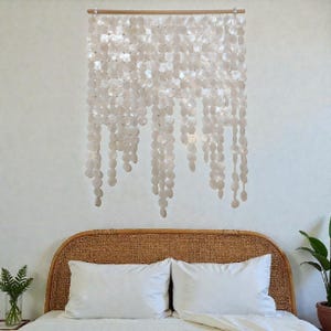 Può includere: Un arazzo decorativo da parete composto da numerosi piccoli dischi di conchiglia rotondi, bianchi. Le conchiglie sono infilate e sospese in linee verticali da un'asta di legno. L'arazzo è sopra un letto con cuscini bianchi e una testiera intrecciata.