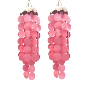 May include: Two pink shell wind chimes. Each chime has a natural-coloured top with hanging strands of pink, round shells. The shells are strung together, creating a cascading effect. The chimes are suspended by thin, silver-coloured strings.
