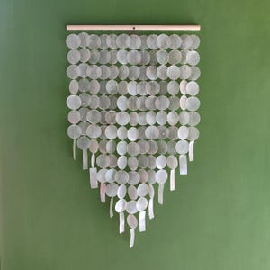 May include: A decorative wall hanging made of numerous iridescent, circular, and rectangular shell pieces. The shell pieces are strung together and suspended from a wooden rod, creating a cascading, triangular shape. The background is a solid green color.