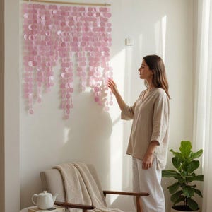 Może przedstawiać: Dekoracja ścienna z różowymi okręgami zawieszonymi na drewnianym drążku. Dzieło sztuki jest zamontowane na białej ścianie, podkreślając estetykę pomieszczenia. Pokój obejmuje również krzesło, stolik boczny i roślinę doniczkową.