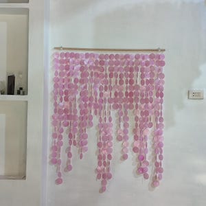 Può includere: Una decorazione murale rosa composta da piccoli pezzi rotondi e iridescenti. La tenda è sospesa a un'asta di legno e pende in fili verticali. Il muro dietro la tenda è bianco.