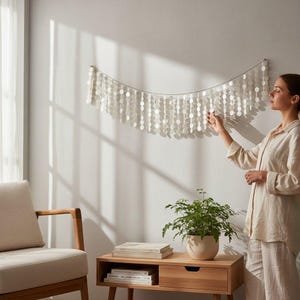 Op de afbeelding: Een wanddecoratie bestaande uit talrijke witte, ronde, schelpachtige schijven die aan een draad hangen. De decoratie is bevestigd aan een witte muur, met een vrouw die het aanpast. Een houten bijzettafel en een plant in een pot staan op de voorgrond.