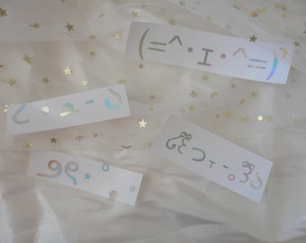 May include: Four white rectangular stickers with holographic designs and symbols. The stickers feature various symbols, including emoticons and decorative elements. The stickers are arranged on a white, star-speckled fabric.