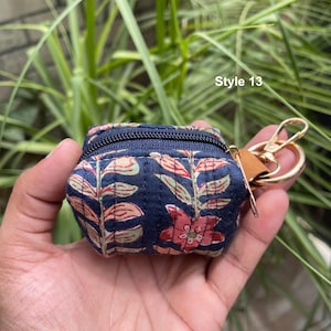 May include: A small, quilted, navy blue zippered pouch featuring a repeating floral and leaf pattern in shades of red, green, and cream. The pouch includes a gold-toned metal lobster clasp keychain attachment and is labeled Style 13.