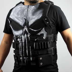 Pode incluir: Colete tático preto com um desenho de caveira branca no peito. O colete tem vários bolsos, correias e um fecho de fivela. O colete é usado sobre uma camiseta preta.