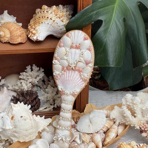 May include: A decorative hairbrush adorned with various seashells, pearls, and coral. The brush features a light pink and white seashell design, with a handle embellished with pearls and a bow. The background includes a wooden shelf with seashells and a large green leaf.