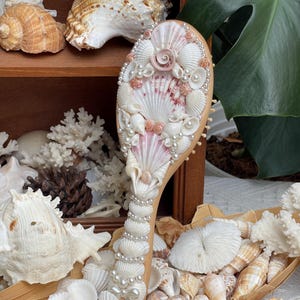 Könnte beinhalten: Eine Holzbürste, verziert mit verschiedenen Muscheln, Perlen und Korallen. Die Bürste hat einen hellen Holzgriff und einen Kopf, der mit weißen, rosa und hellbraunen Muscheln verziert ist. Der Griff ist mit Perlen verziert.