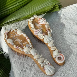 Puede incluir: Dos cepillos de pelo de madera decorados con conchas y perlas. Uno tiene el nombre "Krystal" escrito, y el otro "Mia". Los cepillos están sobre una superficie blanca texturizada.