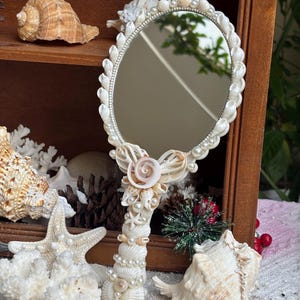 Puede incluir: Un espejo de mano ovalado, intrincadamente decorado con conchas marinas y perlas de imitación. El mango y el marco están adornados con varias conchas, creando un tema costero y náutico. El espejo se presenta contra un fondo de otras conchas y elementos decorativos.