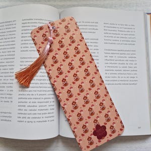 Op de afbeelding: Een roze bladwijzer met een herhalend patroon van kleine beren en aardbeien. De bladwijzer heeft een kwastje en een klein logo met de tekst "Mimose Fairy". De bladwijzer ligt op een open boek.