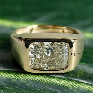 May include: A gold ring with a large, square-cut, light-colored gemstone. The ring has a thick band and a polished finish. The gemstone is set in a bezel setting, and the ring is displayed on a green surface.