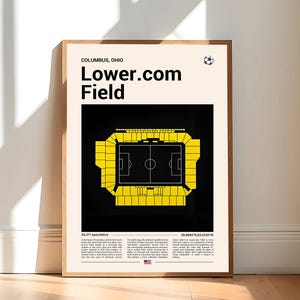 May include: Framed poster featuring a yellow and black illustration of Lower.com Field in Columbus, Ohio. The poster includes the stadium name, a soccer ball icon, and text detailing the stadium's capacity and location. The poster is displayed on a wooden floor.